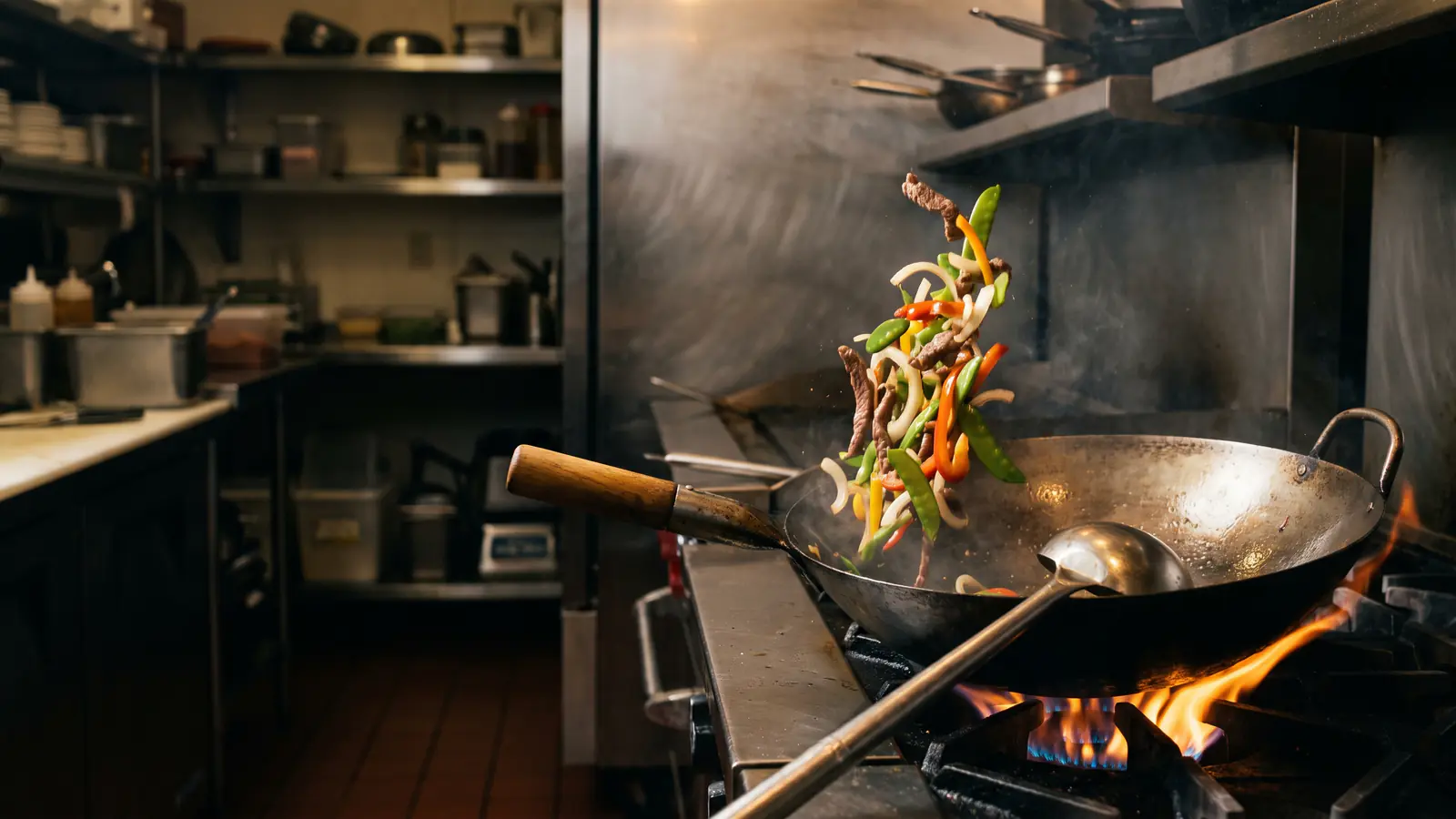 A carbon-steel wok over a gas flame in a dark restaurant kitchen, with pieces of food in the air above the pan in mid-toss and no one in the frame