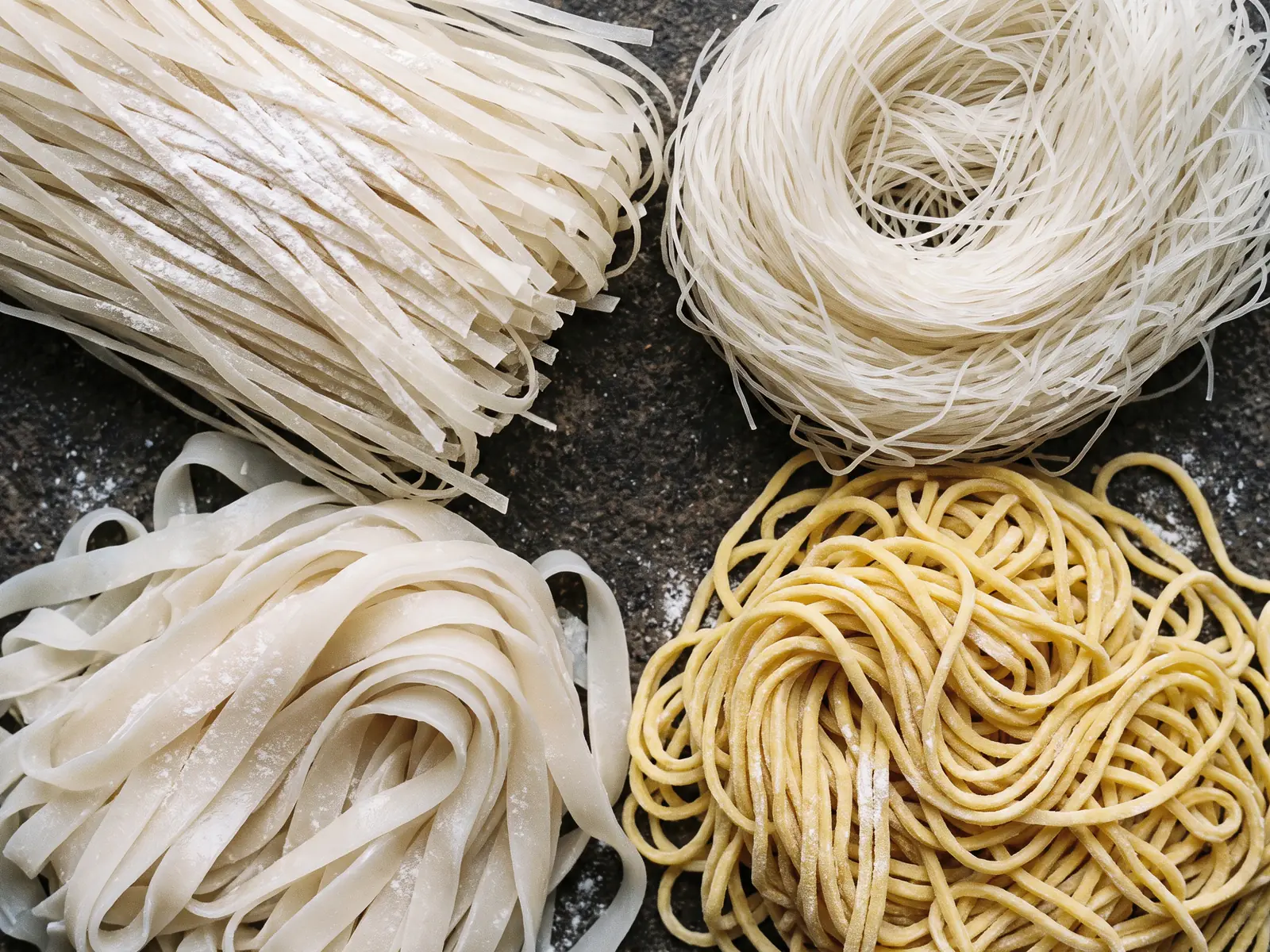 An overhead view of four different kinds of raw noodle laid in separate groups on dark timber, differing in width and colour, with no labels
