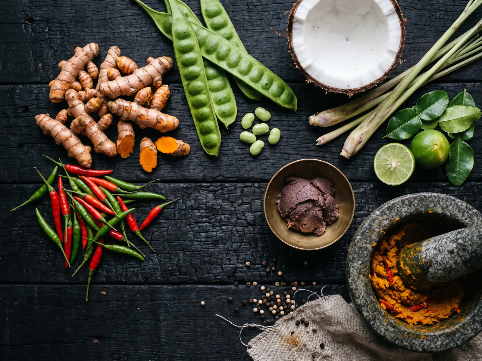 An overhead view on dark wood of fresh turmeric rhizomes snapped open, red and green chillies, flat green sator pods with shelled beans, a halved coconut, lemongrass, lime leaves and lime, a brass bowl of dark shrimp paste and a granite mortar of pounded turmeric paste