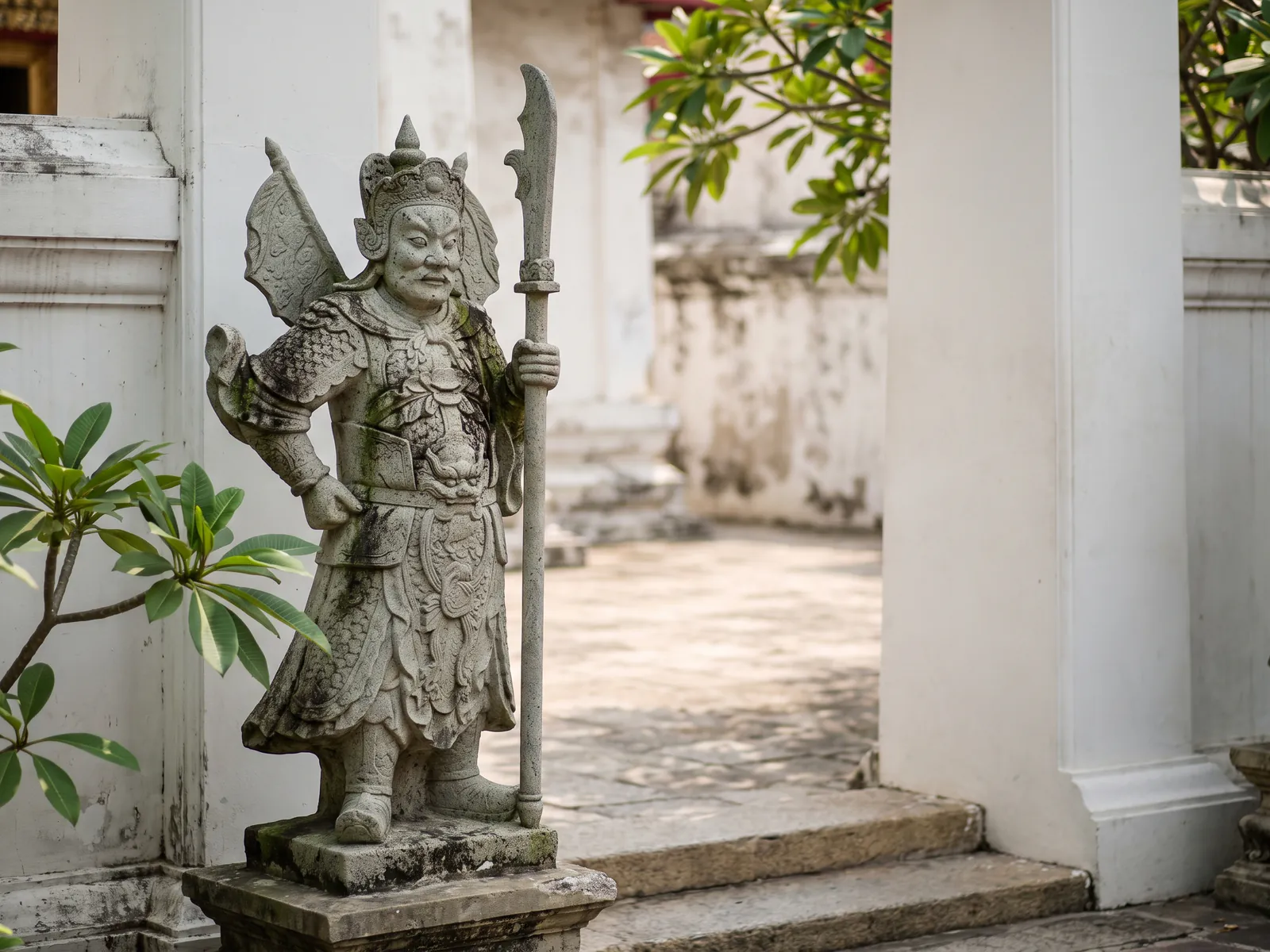 A weathered grey stone statue of a Chinese warrior in armour and a crested helmet, with banners at his back, holding a long-bladed halberd beside a white temple wall