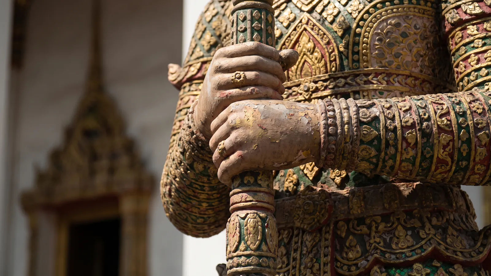 Close view of a temple guardian statue's hands gripping a club, its armour covered in small pieces of glazed porcelain in gold, red and green