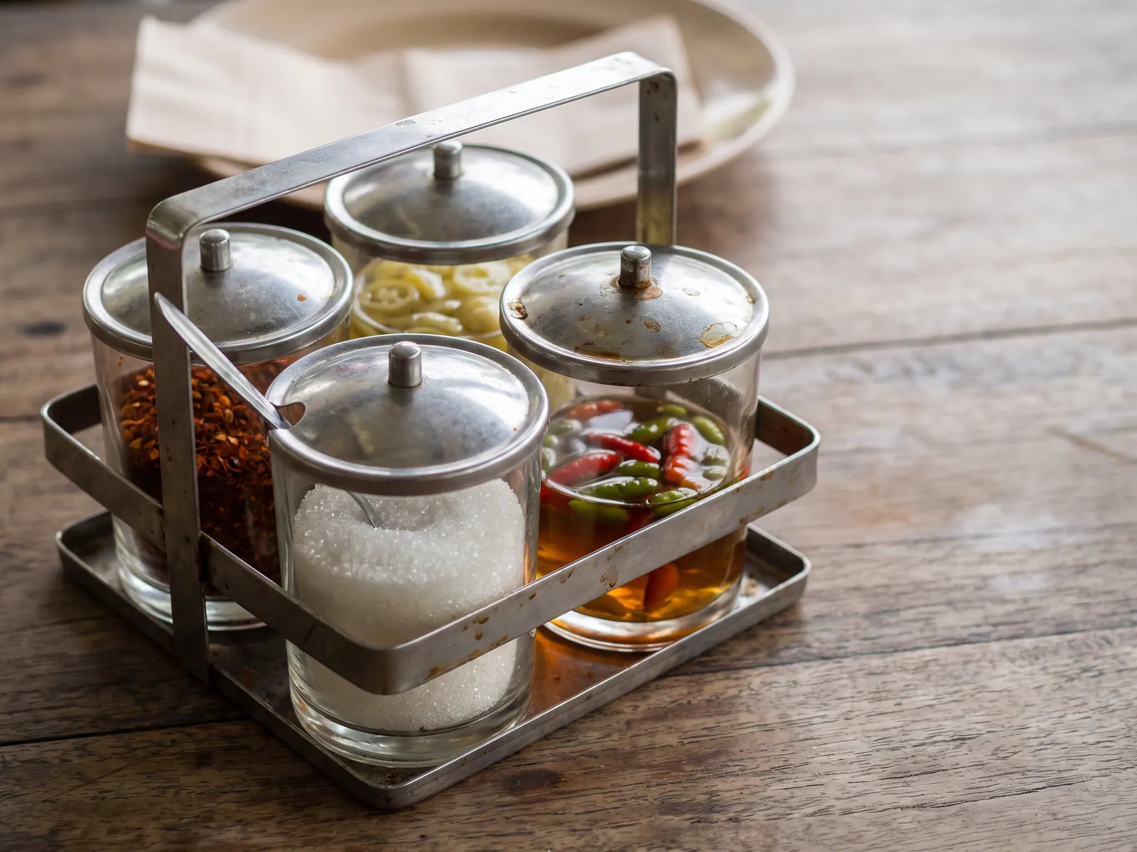 A well-used steel caddy holding four glass pots on a timber table, filled with dried chilli flakes, sugar, sliced green chillies in vinegar and chillies in fish sauce, with small spoons