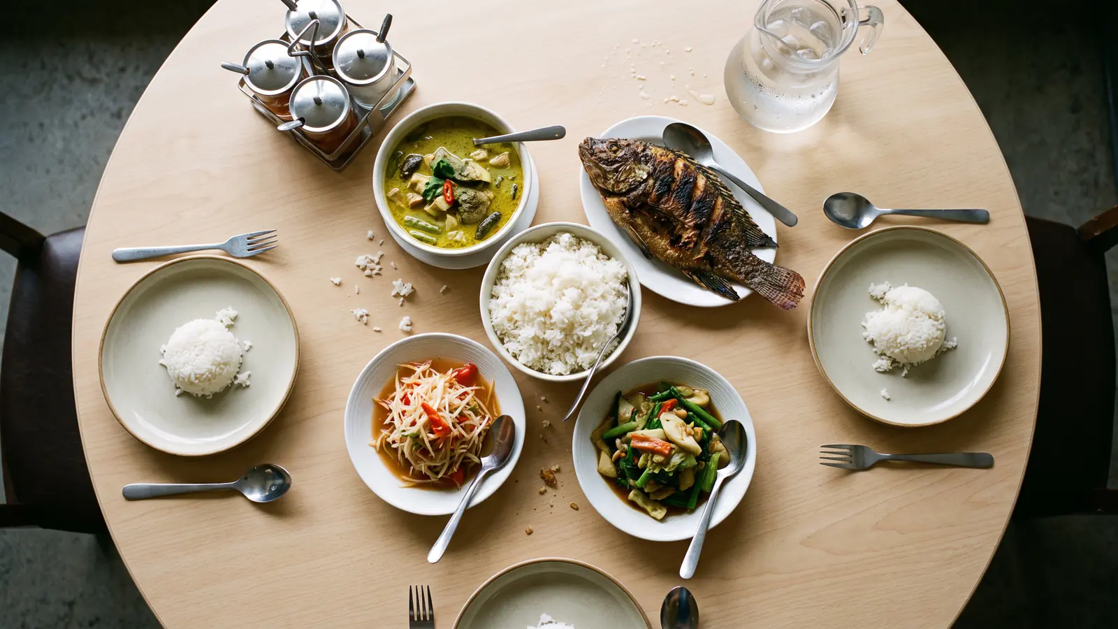 An overhead view of a round pale-wood table mid-meal, with a green curry, a whole grilled fish, a papaya salad and stir-fried greens in the middle, a bowl of steamed rice at the centre, and individual plates of rice with a fork and spoon around the edge