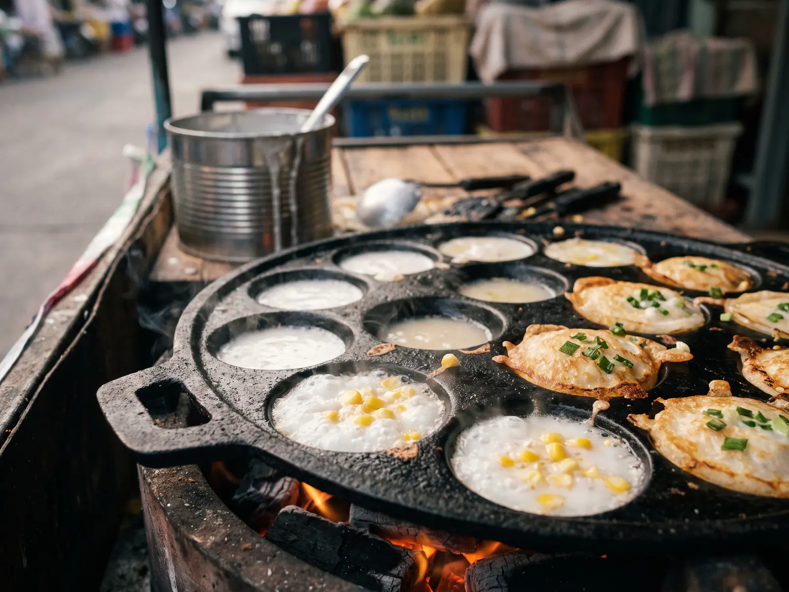 A cast-iron pan of coconut pancakes over glowing charcoal, the wells at different stages: some just filled with pale batter, others set with a golden edge and a pool of coconut cream